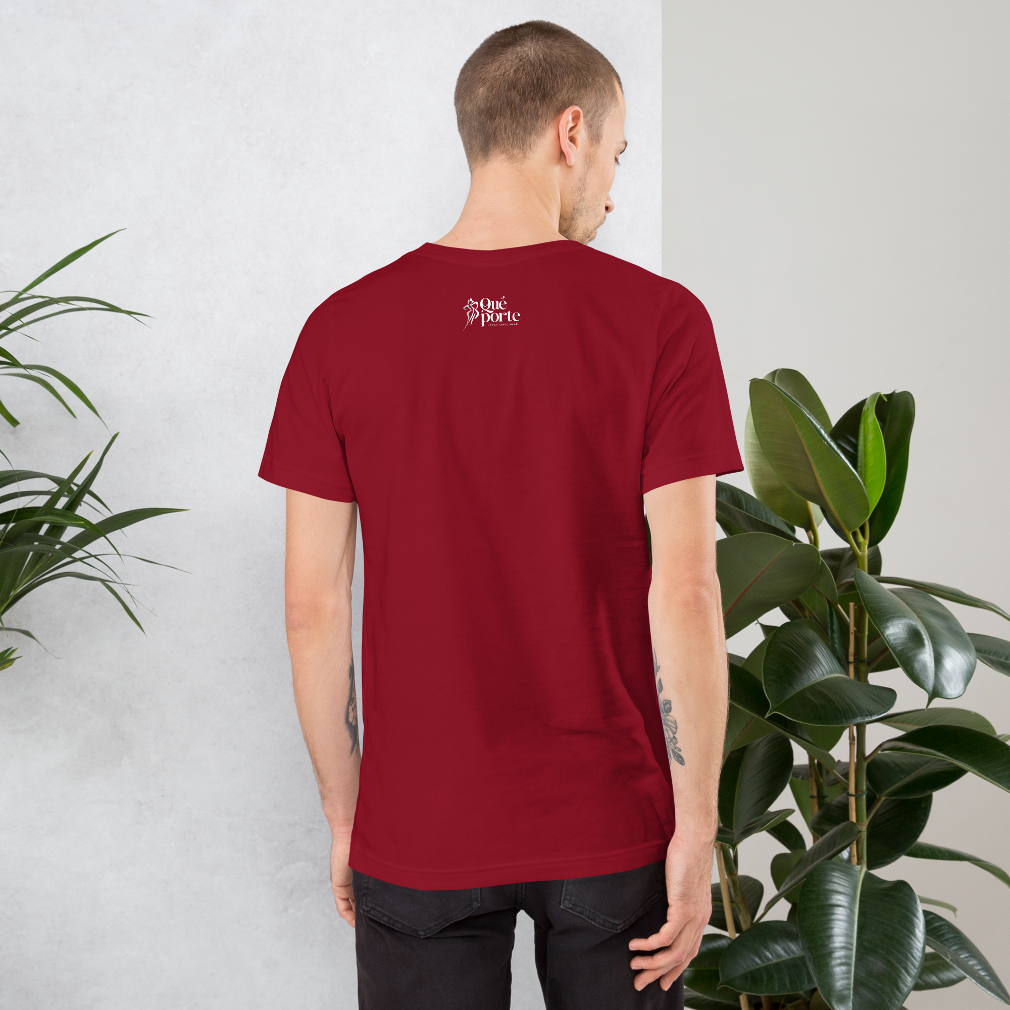 Man wearing a red t-shirt with a logo on the back, standing against a light gray wall with plants.
