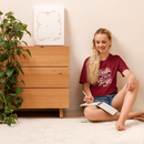 Woman sitting on the floor reading a book in a room with a wooden dresser and plant.