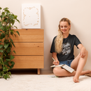 Woman sitting on the floor reading a book in a room with a wooden dresser and plant.