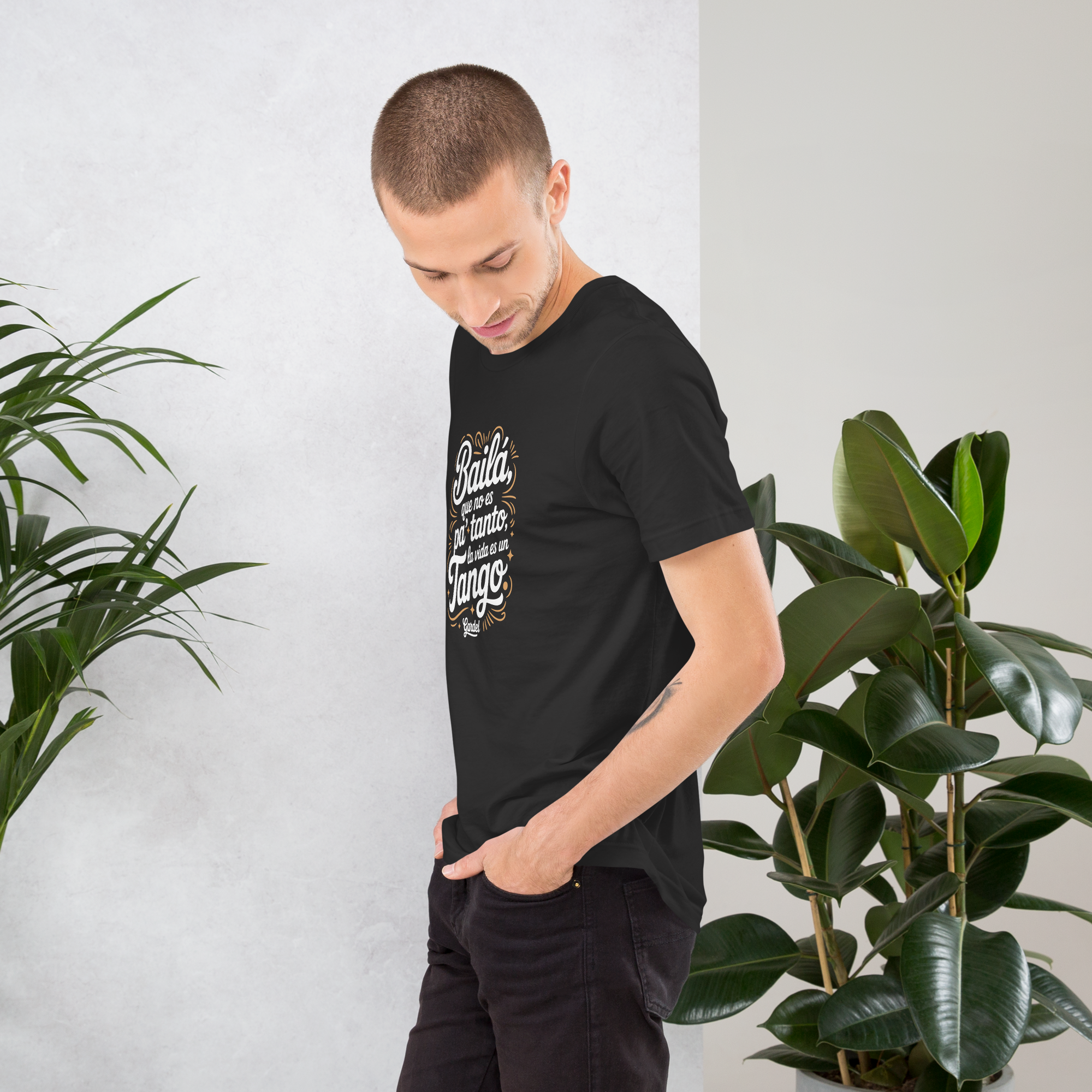 Man wearing a black t-shirt with text, standing between two potted plants against a white wall.