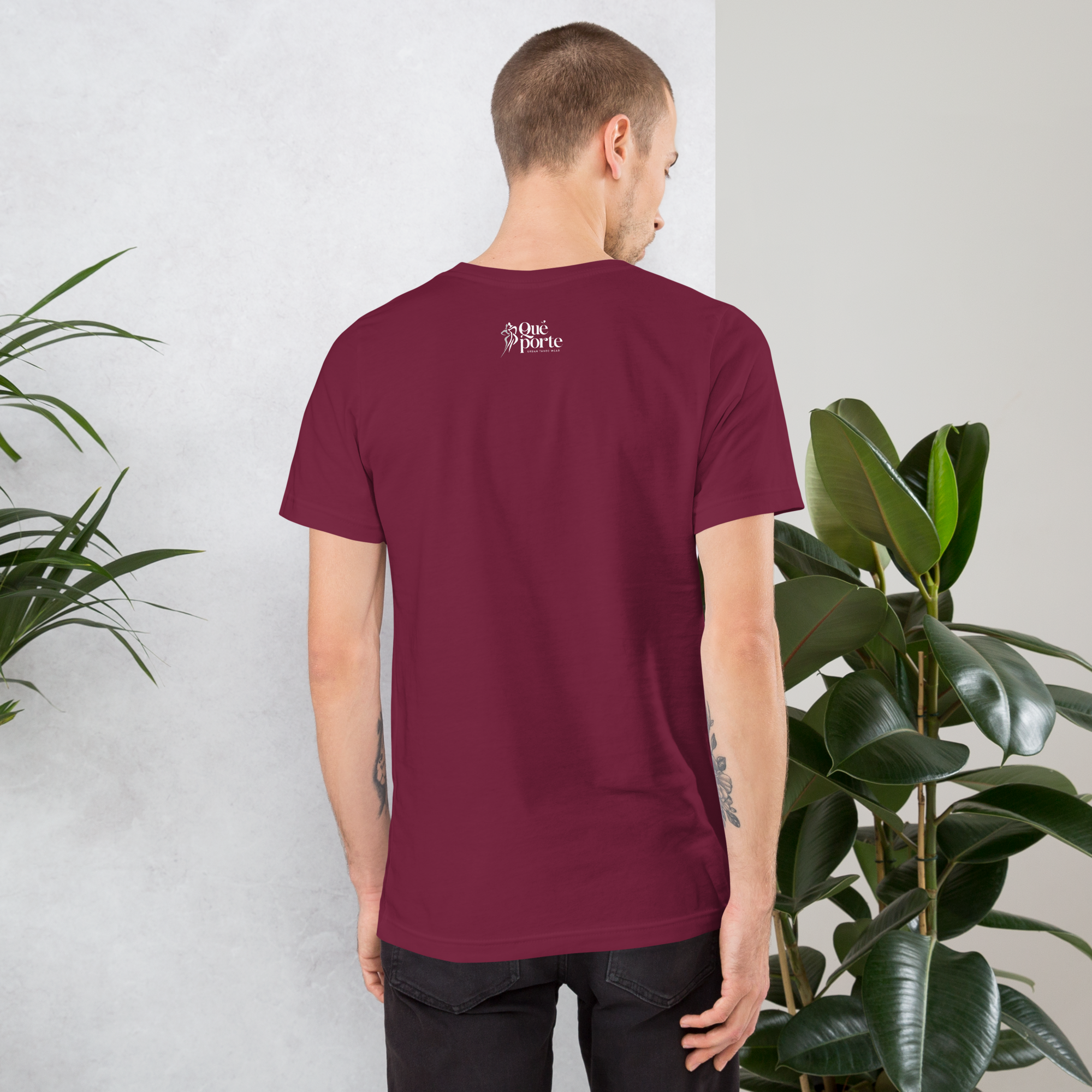 Person wearing a maroon t-shirt with a logo on the back, standing in front of a white wall with plants.