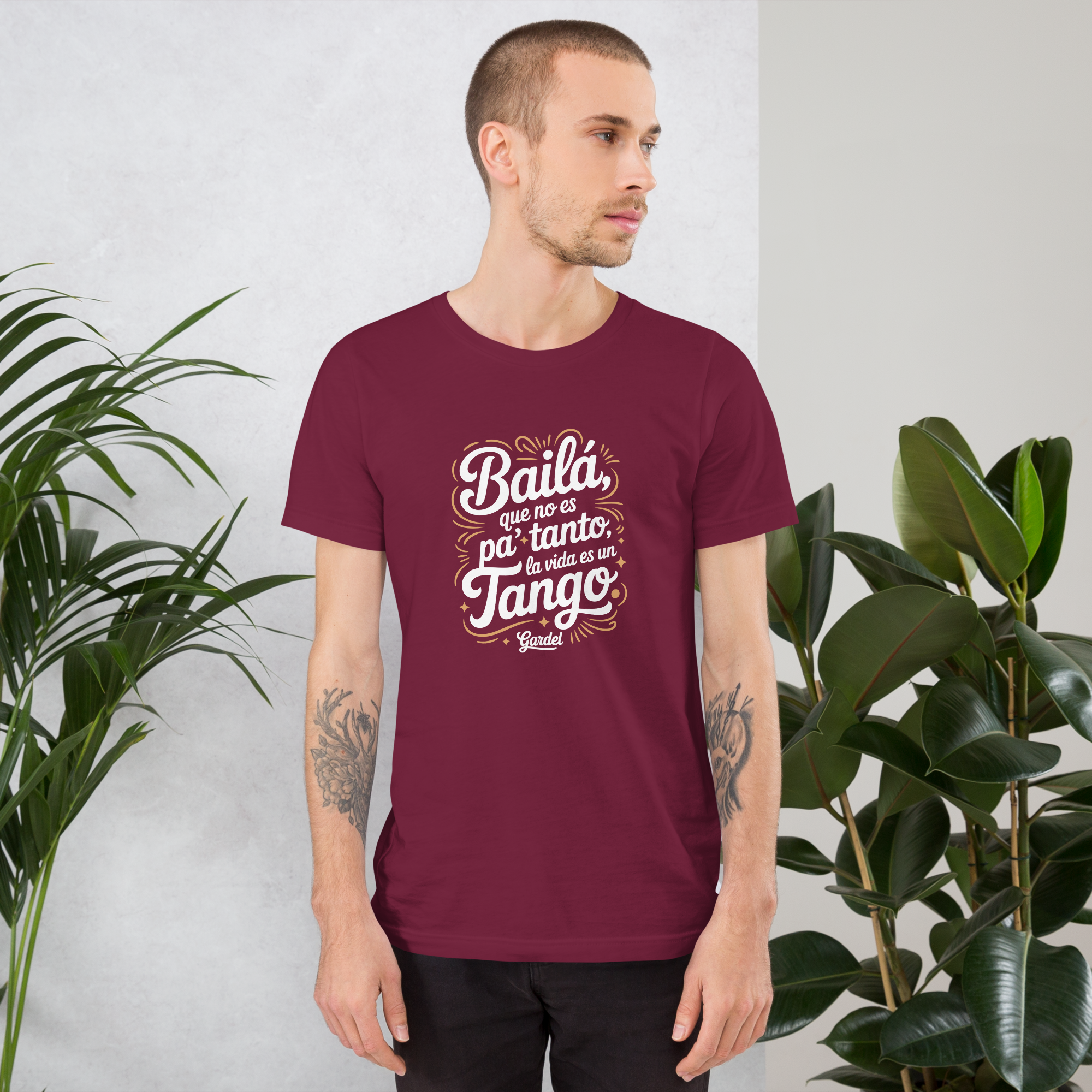 Man wearing a maroon t-shirt with Spanish text, standing against a light gray background with plants.
