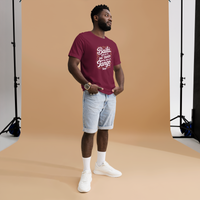 Man wearing a maroon t-shirt with text and light blue shorts standing against a brown backdrop.