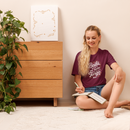 Woman sitting on the floor reading a book in a room with a wooden dresser and plant.