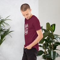 Man wearing a maroon t-shirt with text, standing between two potted plants against a white wall.