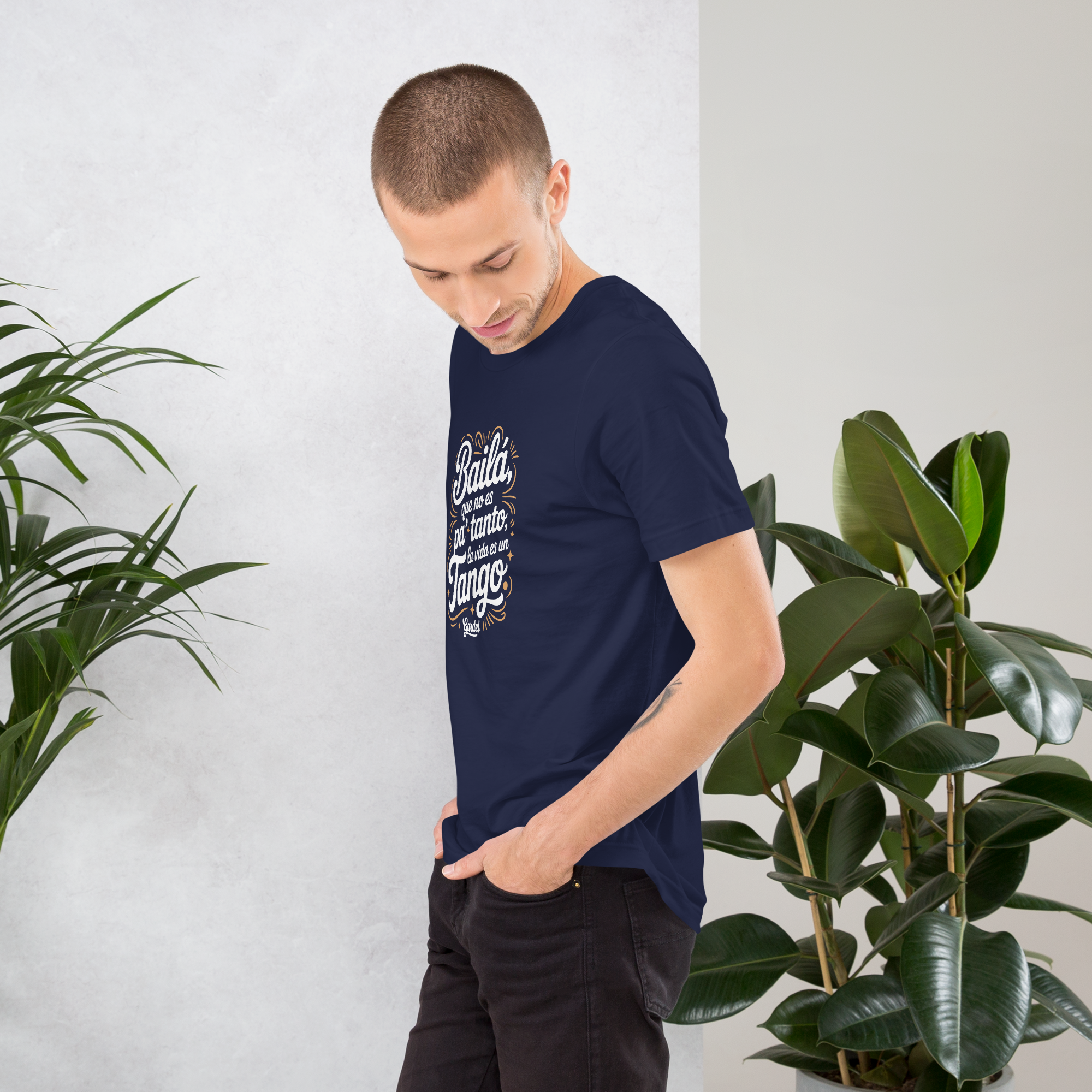 Man wearing a navy blue t-shirt with text, standing between two potted plants against a white wall.