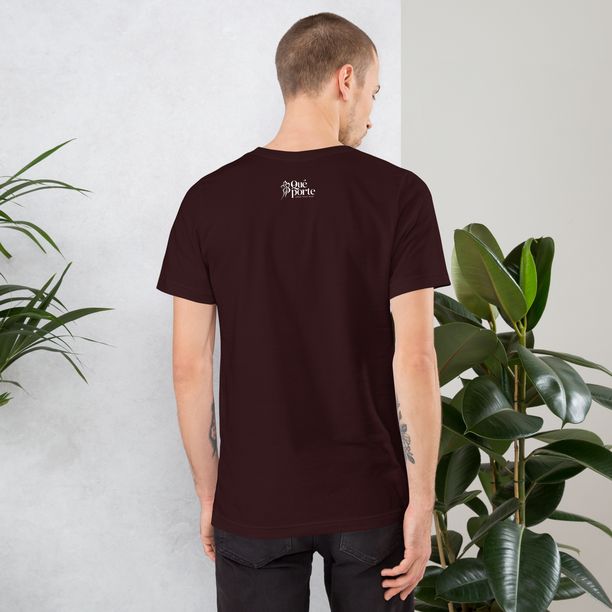 Man wearing a dark brown t-shirt with a logo on the back, standing against a light gray wall with plants.