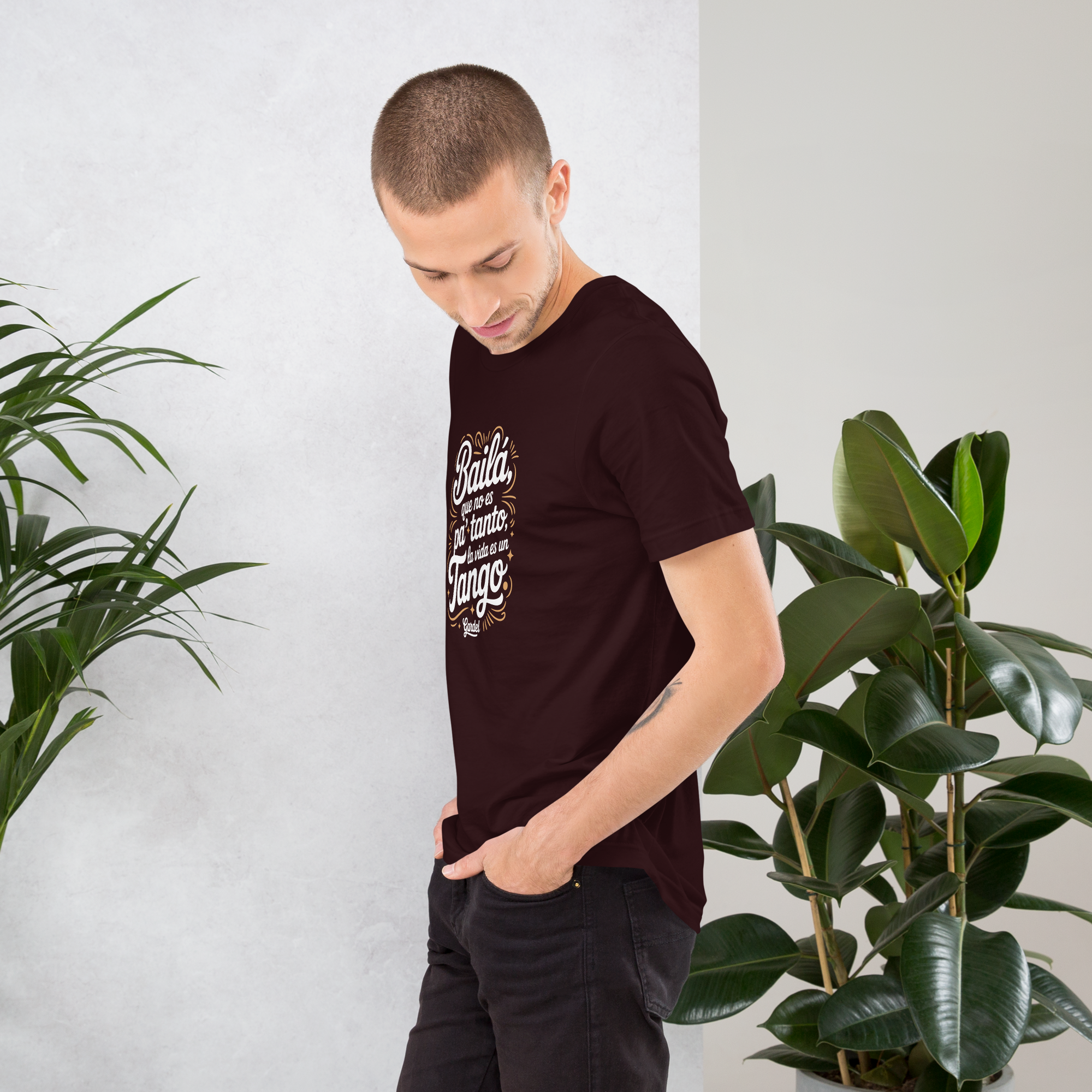 Man wearing a dark t-shirt with text, standing between two potted plants against a white wall.