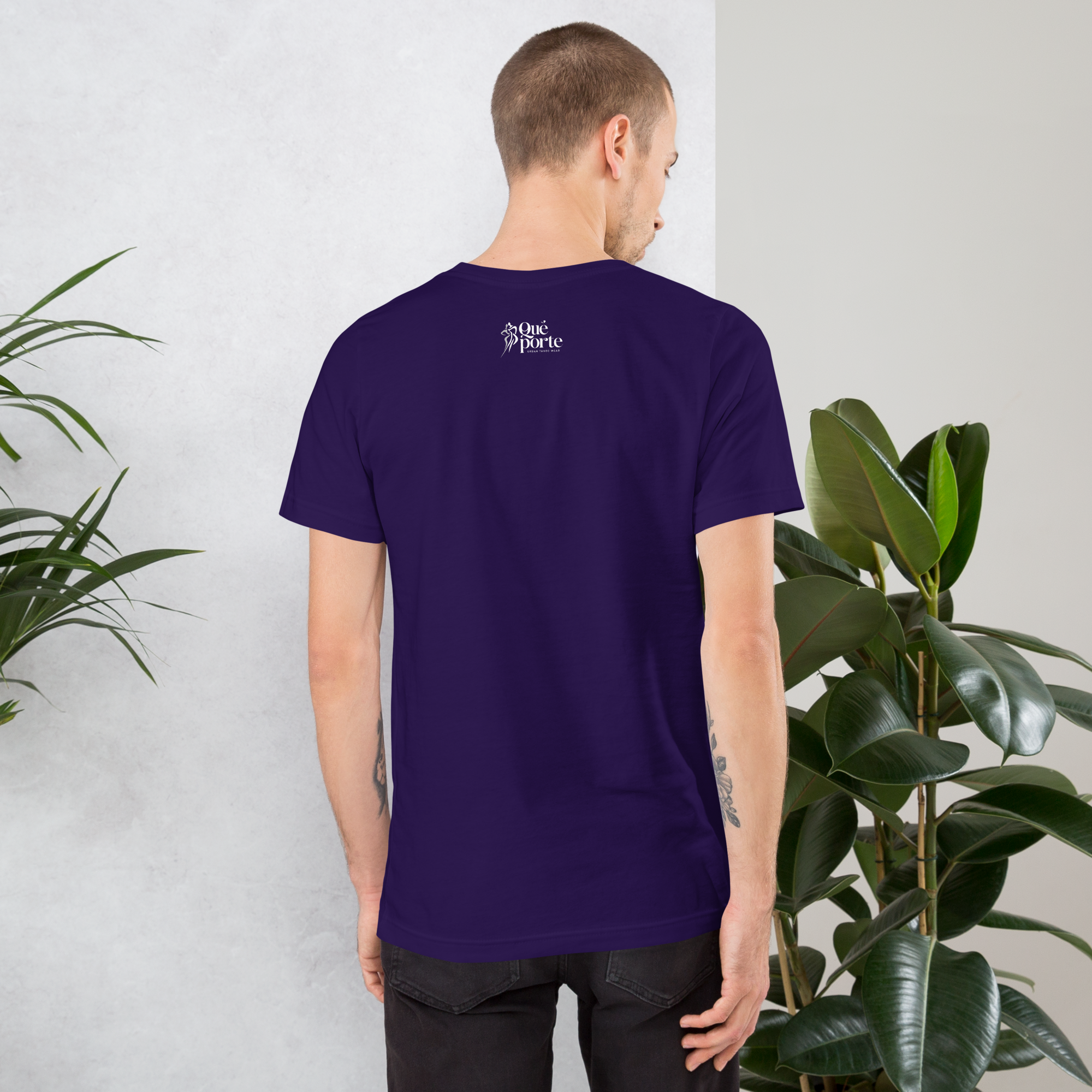 Man wearing a purple t-shirt with text on the back, standing against a light gray wall with plants.