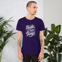 Man wearing a purple t-shirt with text standing between two plants against a light gray wall.