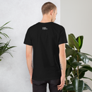 Man wearing a black t-shirt with a logo on the back, standing against a light gray wall with plants.