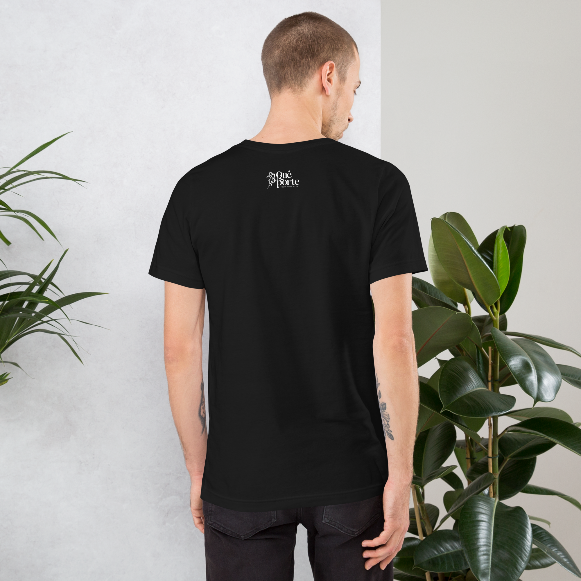 Man wearing a black t-shirt with a logo on the back, standing against a light gray wall with plants.